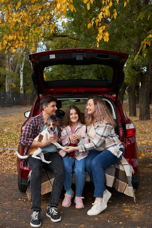 Happy parents with daughter and cute dog Jack Russel terrier sitting in car trunk on autumn day. Long auto journey break.の写真素材