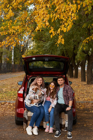 Happy parents with daughter and cute dog Jack Russel terrier sitting in car trunk on autumn day. Long auto journey break.の写真素材