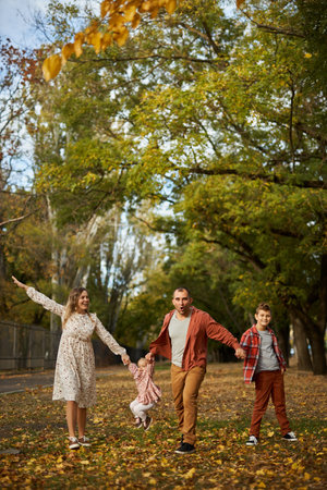 Happy family walking on green grass strewn with fallen leaves in Park in autumn. Parents, two children are walking in park at sunset with holding hands. Family, child, walk through Autumn Park.の写真素材