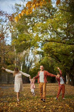 Happy family walking on green grass strewn with fallen leaves in Park in autumn. Parents, two children are walking in park at sunset with holding hands. Family, child, walk through Autumn Park.の写真素材