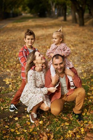 Happy family walking on green grass strewn with fallen leaves in Park in autumn. Parents, two children are walking in park hug and laugh. Family, child, walk through Autumn Park.の写真素材
