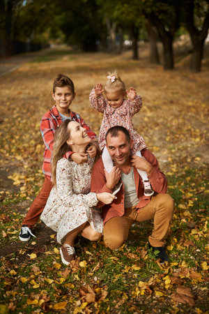 Happy family walking on green grass strewn with fallen leaves in Park in autumn. Parents, two children are walking in park hug and laugh. Family, child, walk through Autumn Park.の写真素材