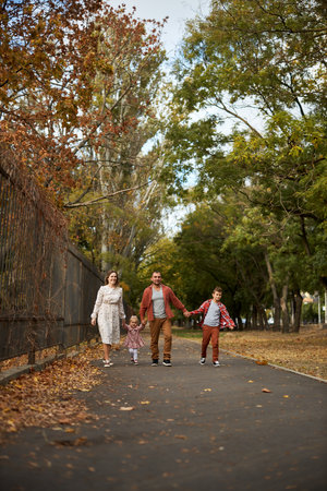 Happy family walking on green grass strewn with fallen leaves in Park in autumn. Parents, two children are walking in park at sunset with holding hands. Family, child, walk through Autumn Park.の写真素材