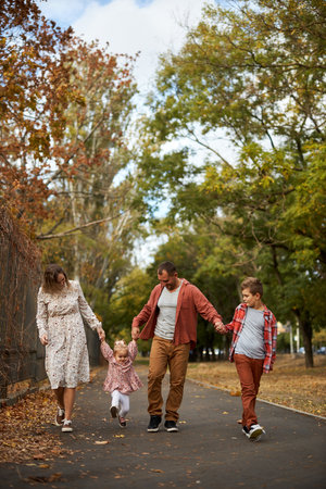 Happy family walking on green grass strewn with fallen leaves in Park in autumn. Parents, two children are walking in park at sunset with holding hands. Family, child, walk through Autumn Park.の写真素材