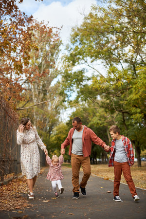 Happy family walking on green grass strewn with fallen leaves in Park in autumn. Parents, two children are walking in park at sunset with holding hands. Family, child, walk through Park.の写真素材