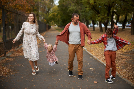 Happy family walking on green grass strewn with fallen leaves in Park in autumn. Parents, two children are walking in park at sunset with holding hands. Family, child, walk through Autumn Park.の写真素材