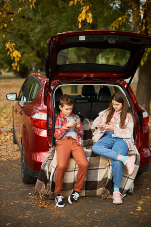 Two children sit in the trunk of a car and use smartphones, play games or surf the internet. Family trip in autumnの写真素材