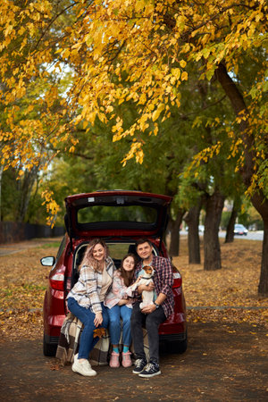 Happy parents with daughter and cute dog Jack Russel terrier sitting in car trunk on autumn day. Long auto journey break.の写真素材