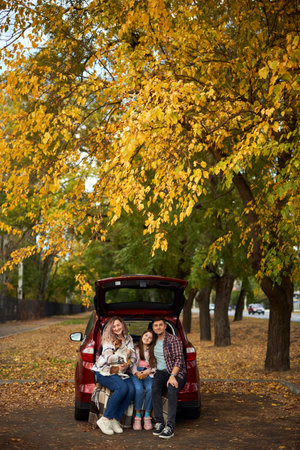 Happy parents with daughter and cute dog Jack Russel terrier sitting in car trunk on autumn day. Long auto journey break.の写真素材