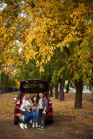 Happy parents with daughter and cute dog sitting in car trunk on autumn day. Long auto journey break.の写真素材