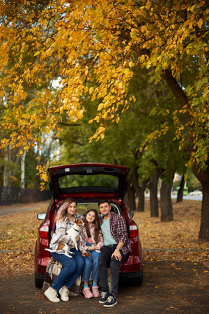 Happy parents with daughter and cute dog Jack Russel terrier sitting in car trunk on autumn day. Long auto journey break.の写真素材