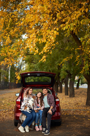 Happy parents with daughter and cute dog Jack Russel terrier sitting in car trunk on autumn day. Long auto journey break.の写真素材