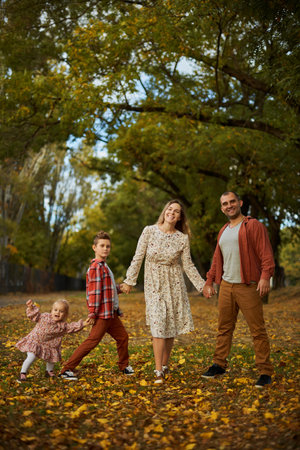 Happy family walking on green grass strewn with fallen leaves in Park in autumn. Parents, two children are walking in park at sunset with holding hands. Family, child, walk through Autumn Park.の写真素材