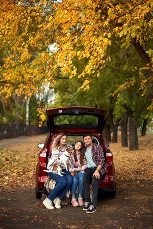 Happy parents with daughter and cute dog Jack Russel terrier sitting in car trunk on autumn day. Long auto journey break.の写真素材