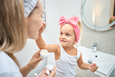 Smiling young woman and little daughter with a cosmetic bow on their heads smearing face cream on faces, during skincare procedure in the bathroom at homeの写真素材