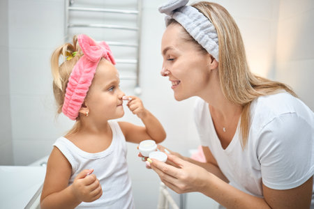 Smiling young woman and little daughter with a cosmetic bow on their heads smearing face cream on faces, during skincare procedure in the bathroom at homeの写真素材