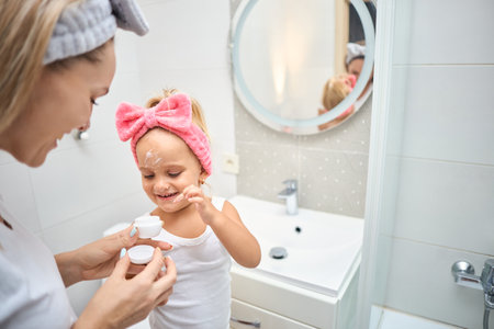 Smiling young woman and little daughter with a cosmetic bow on their heads smearing face cream on faces, during skincare procedure in the bathroom at homeの写真素材