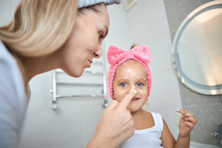 Smiling young woman and little daughter with a cosmetic bow on their heads smearing face cream on faces, during skincare procedure in the bathroom at homeの写真素材
