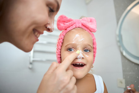 Smiling young woman and little daughter with a cosmetic bow on their heads smearing face cream on faces, during skincare procedure in the bathroom at homeの写真素材