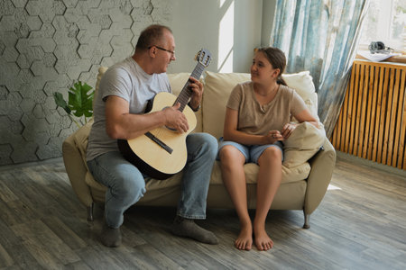 Grandfather and granddaughter are in the room on the sofa, he plays the guitar and sings. Spending time together, family, old ageの写真素材