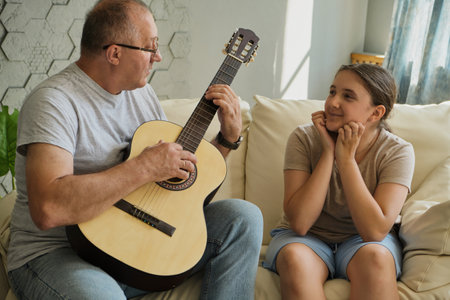 Grandfather and granddaughter are in the room on the sofa, he plays the guitar and sings. Spending time together, family, old ageの写真素材