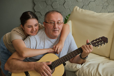 Kid girl hugging her grandfather as he plays guitar. Spending time together, family, old ageの写真素材