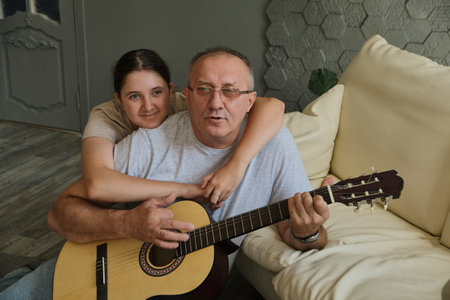 Kid girl hugging her grandfather as he plays guitar. Spending time together, family, old ageの写真素材