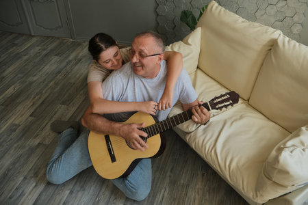 Kid girl hugging her grandfather as he plays guitar. Spending time together, family, old ageの写真素材