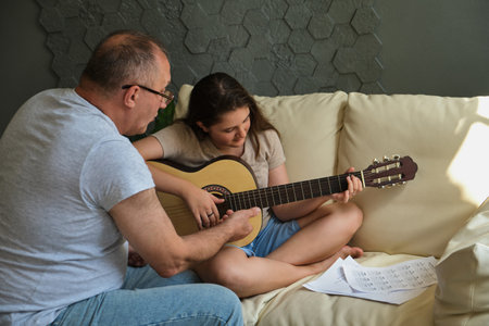 Grandfather teaching his granddaughter to play guitar on sofa at home. Spending time together, family, old ageの写真素材