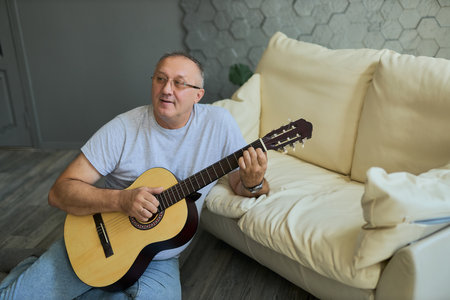 Senior Man playing acoustic guitar at home, relaxing on the floor next to a sofa. Ideal for music, hobby, and leisure themesの写真素材