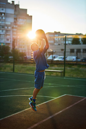 Sports and basketball. Boy in a blue tracksuit throwing a basketball ball into the hoop while playing on an outdoor court In the rays of the sunset. Sports conceptの写真素材