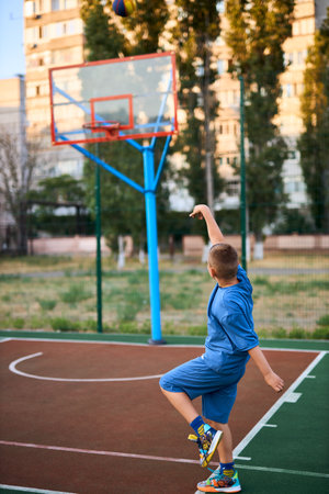 Sports and basketball. Boy in a blue tracksuit throwing a basketball ball into the hoop while playing on an outdoor court. Residential urban area in the background. Sports conceptの写真素材