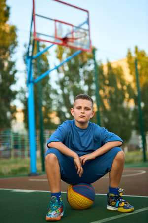 Young tired sporty boy after training wearing blue sportswear looking at camera, sitting on the ball at basketball game playground court. Outdoor courtyard sports concept.の写真素材