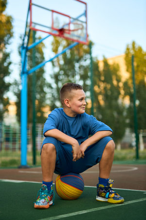Young tired sporty boy after training wearing blue sportswear looking aside sitting on the ball at basketball game playground court. Outdoor courtyard sports concept.の写真素材