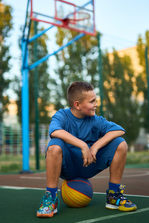 Young tired sporty boy after training wearing blue sportswear looking aside sitting on the ball at basketball game playground court. Outdoor courtyard sports concept.の写真素材