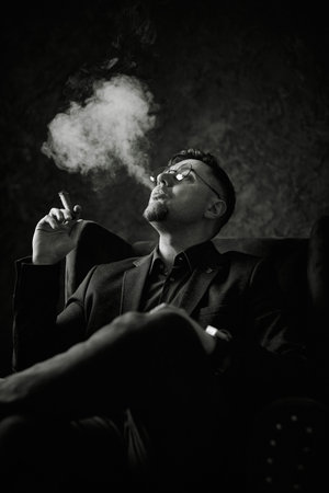 Monochromatic portrait of a serious man, wearing a suit jacket and sitting smoking a cigar. Smoke swirls around him against a dark background, creating a sophisticated, contemplative or powerful moodの写真素材
