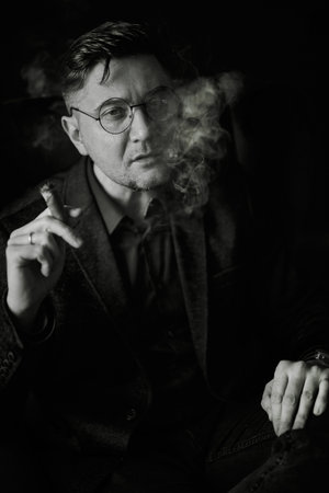 Monochromatic portrait of a serious man, wearing a suit jacket and sitting smoking a cigar. Smoke...