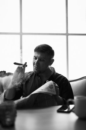 World economy political news concept. Luxury thoughtful chic dandy man, owner reading morning newspaper smoking a cigar while sitting indoors near a window with panes. Monochromatic portraitの写真素材