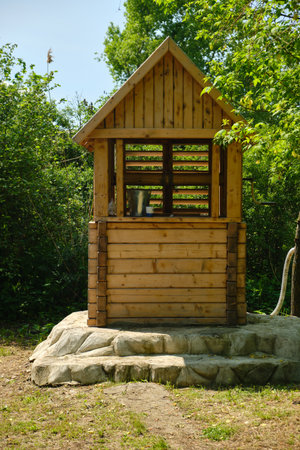 rustic wooden water well with a bucket inside stands on stone steps surrounded by vibrant green foliage and trees on a sunny day. Concept of rural life and natural resourcesの写真素材