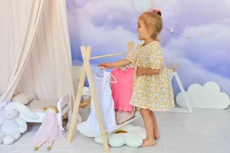 cute baby girl in a peach dress stands by a wooden clothes rack, looking at garments choosing what to wear, playful room. childhood, play, and domestic lifeの写真素材