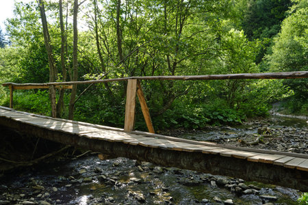 A simple handmade wooden footbridge crosses a shallow rocky river in a lush summer forest. Sunlight filters through the trees, creating a peaceful scene of rural travel and natureの写真素材