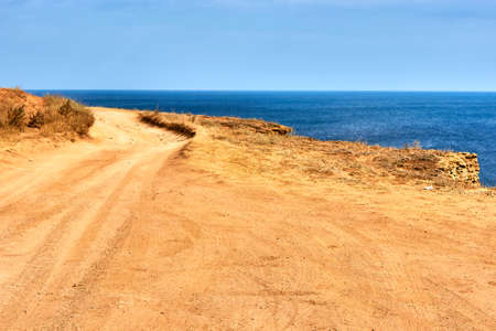 A sandy country road along the edge of the cliff. Orange land, a cape by the sea. Seascape, horizon, calm blue sea, clear sky.の写真素材