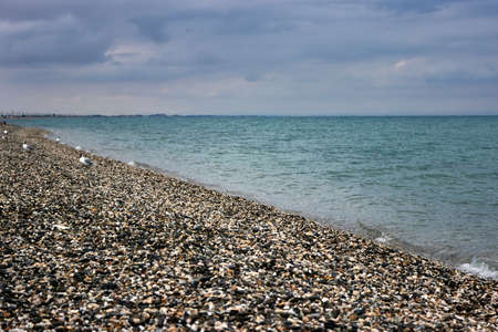 Calm blue sea in the evening. View of the water surface from the pebble beach. Gulls go into the distance along the shore. Dark clouds in the sky. The coast on the diagonal photo. Natural marine landscape.の写真素材