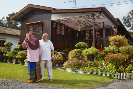 Senior couple smiling and waving in front of wooden houseの写真素材