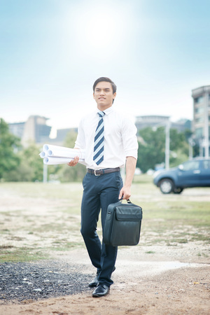 Young businessman holding briefcase and blueprints on siteの写真素材