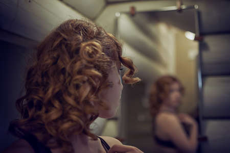 Curly hair redheaded young woman looking at herself in the mirror in the bathroom