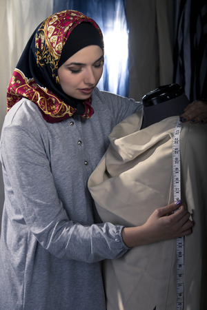 Female fashion designer tailoring conservative clothing in a textile workshop.  The hijab she is wearing is associated with muslims, middle east or eastern european cultures.  The image depicts creativity and professional seamstress craft.の写真素材