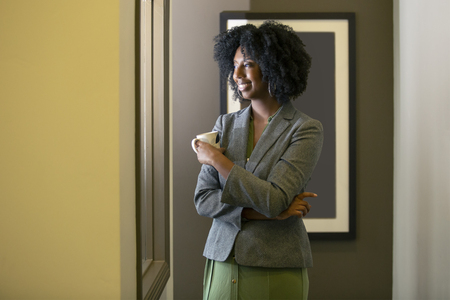 Black African American businesswoman having coffee in the morning while looking out the office window. The woman looks like she is about to start work or is taking a break. の写真素材