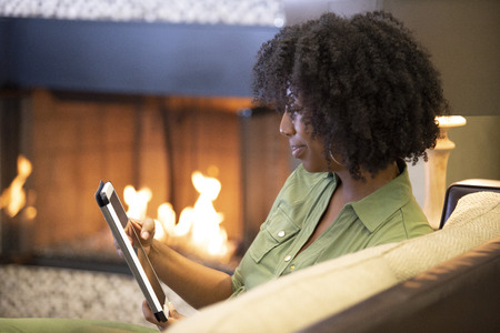 Black African American female relaxing at home and reading or browsing the internet with a digital tablet in a living room by the fireplace.  The image depicts technology in domestic lifestyle.の写真素材