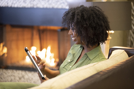 Black African American female relaxing at home and reading or browsing the internet with a digital tablet in a living room by the fireplace.  The image depicts technology in domestic lifestyle.の写真素材